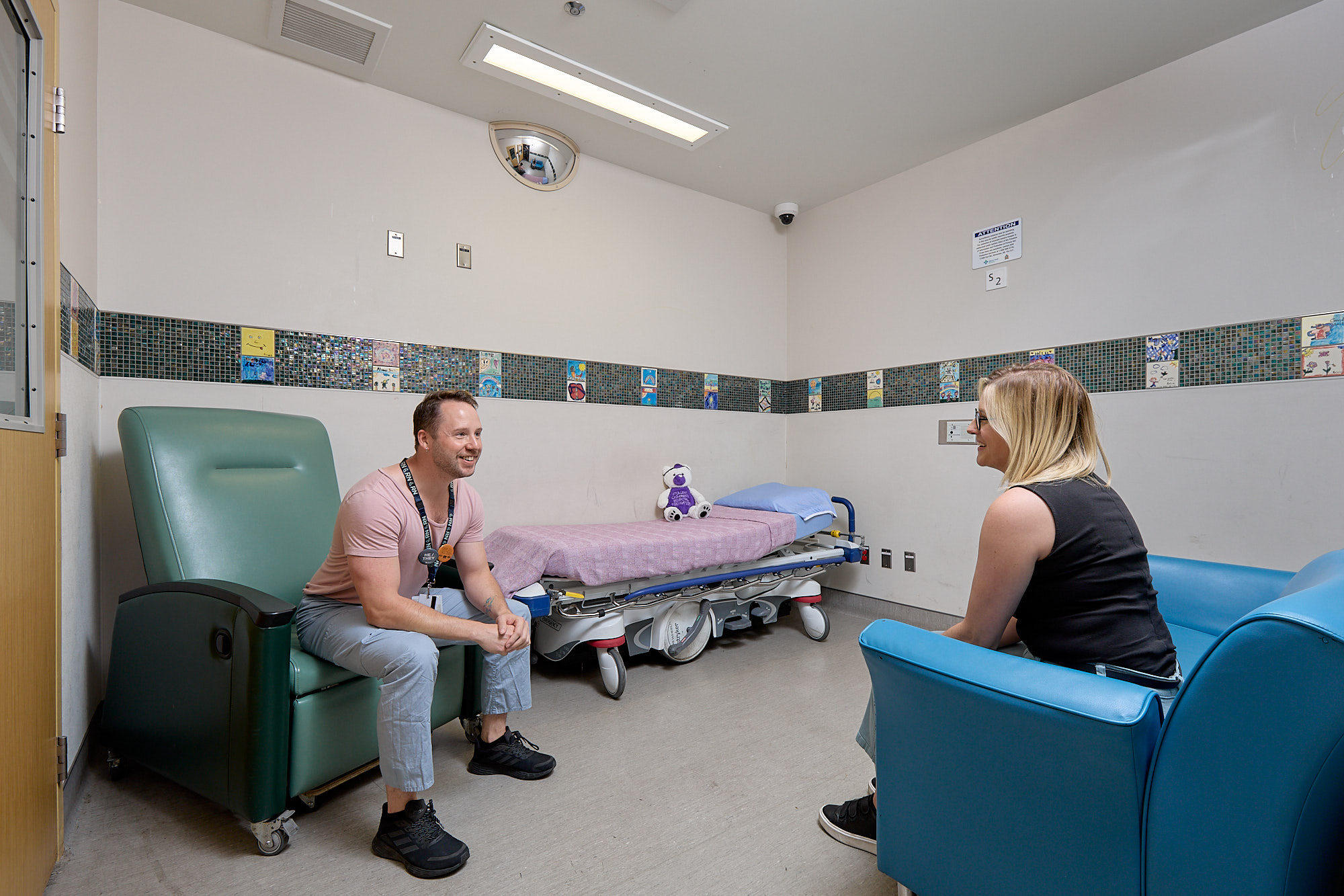 A male mental health medical professional in a pink shirt and scrubs sits in a green chair, supporting teenage patient sitting in a blue chair.
