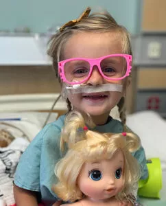 Stollery kid Lukah sitting in hospital bed wearing pink glasses and holding a doll